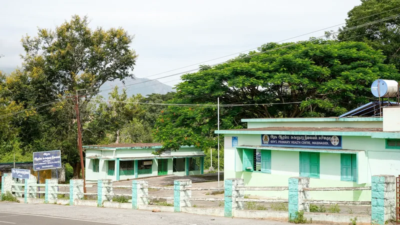 A single-storey government primary health centre in Tamil Nadu, pale green walls and a board in Tamil and English, empty road in front of it.